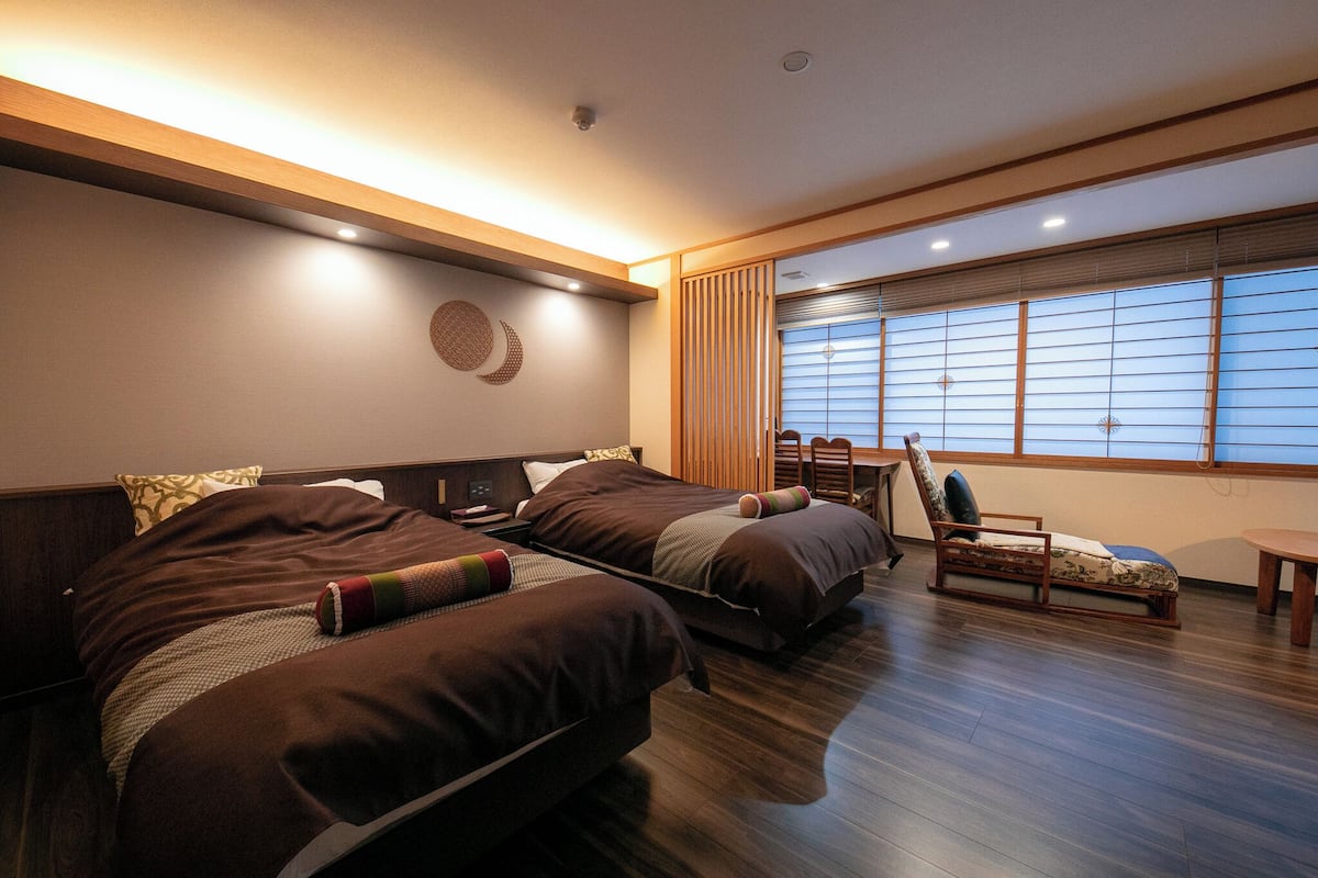 Ein Hotelzimmer mit zwei Betten, Holzboden, einem Fenster mit Jalousien und einem kleinen Tisch.