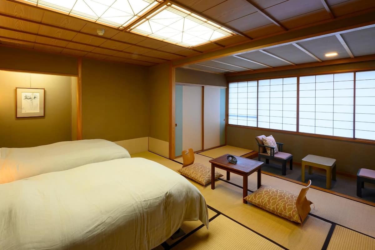 Habitación de hotel con dos camas, una zona de estar con una mesa y sillas, y un ventanal grande.