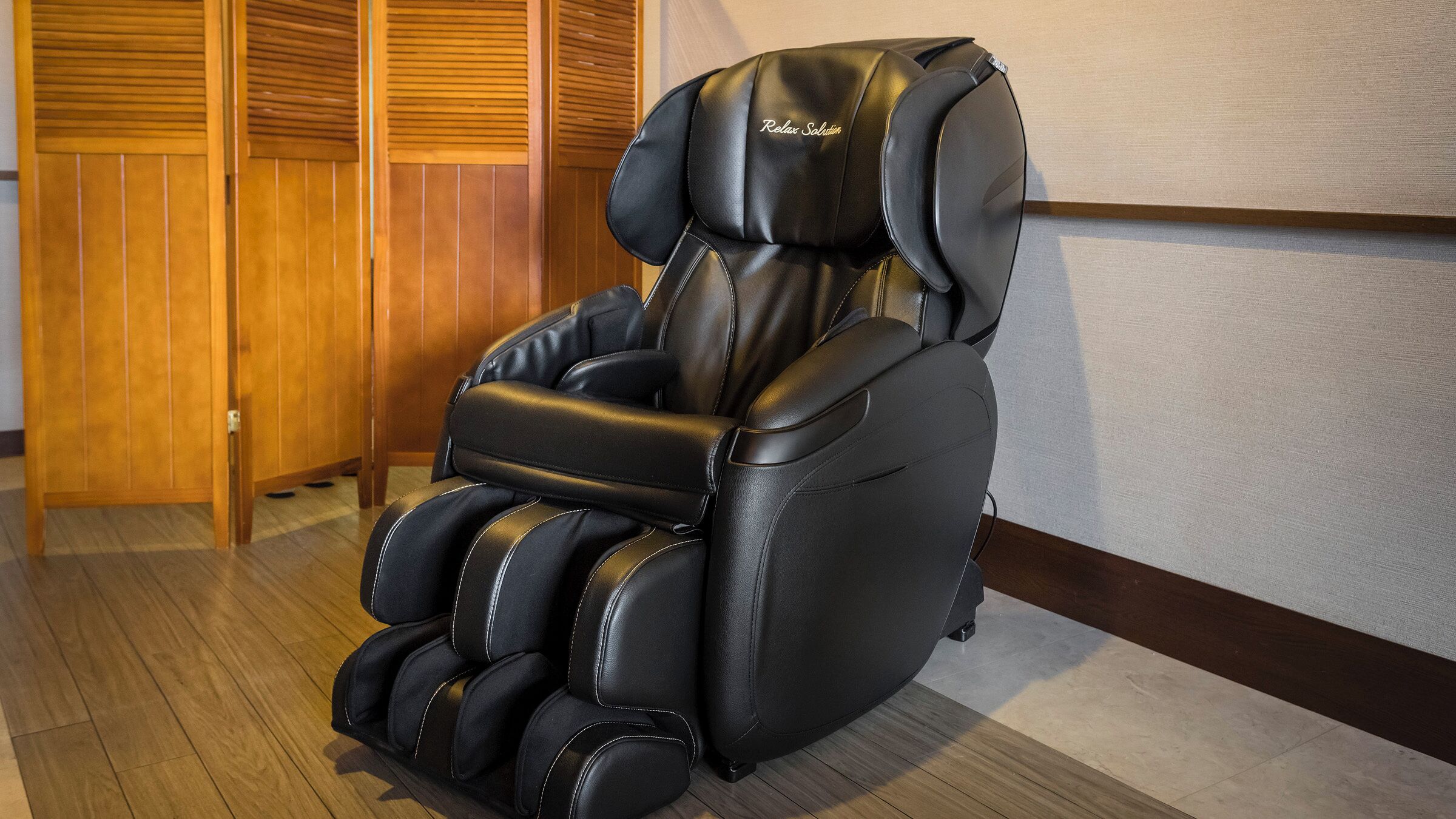 A massage chair with a headrest and footrest, positioned in a room with wooden flooring and a wooden door in the background.