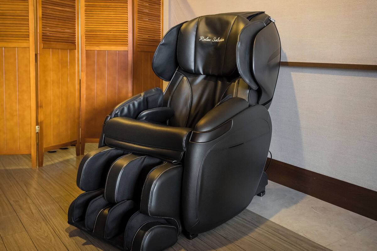 Un fauteuil de massage doté d’un repose-tête et d’un repose-pieds, placé dans une pièce dont le sol est en bois et où l’on aperçoit une porte en bois à l’arrière-plan.