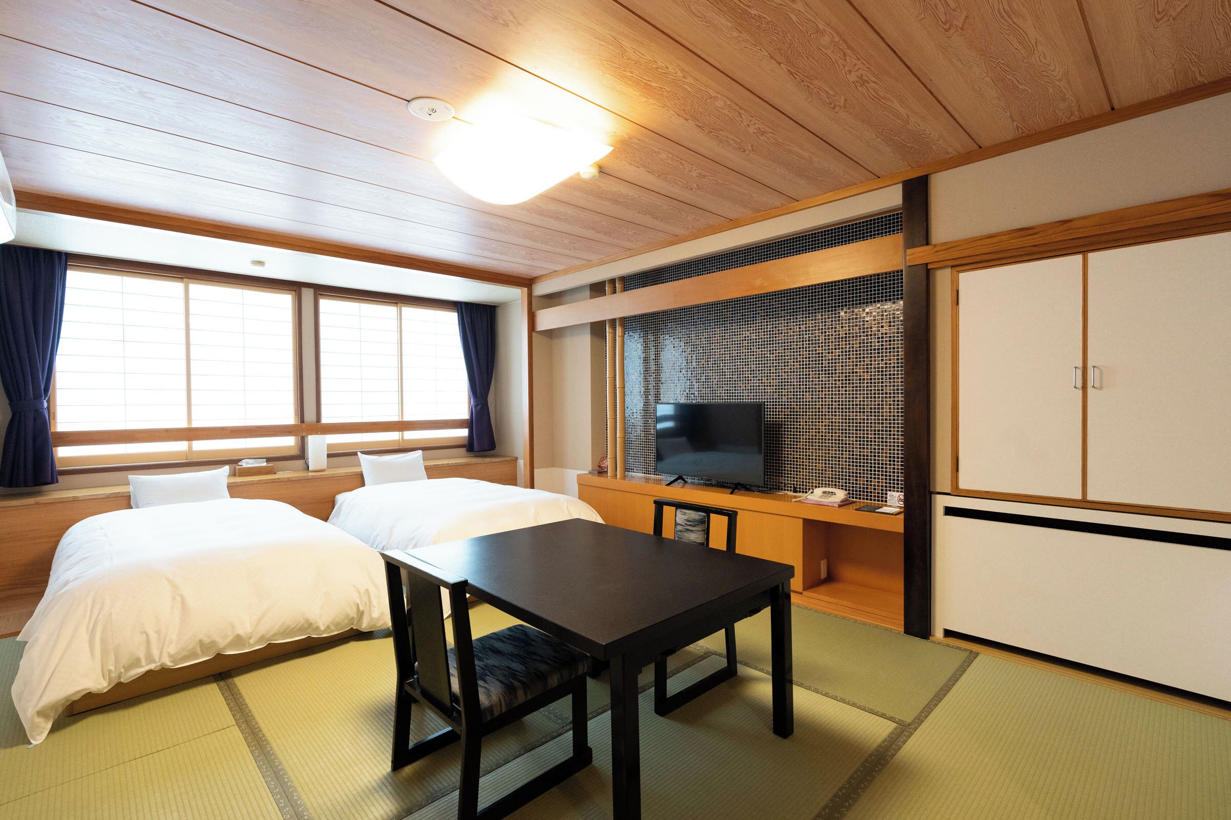Ein traditioneller japanischer Raum mit Tatami-Bodenbelag, einem Bett, einem Schreibtisch und einem Fernseher.