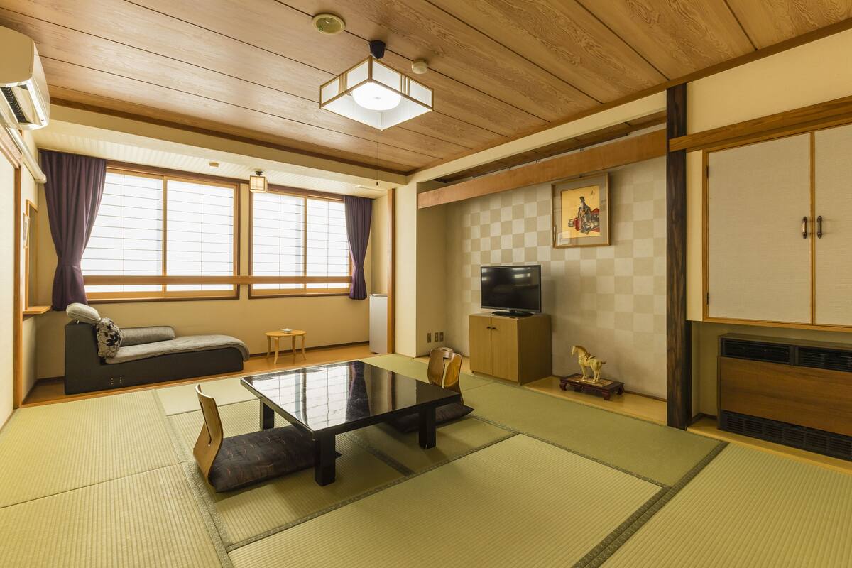 Une pièce de style japonais traditionnel, avec un sol recouvert de tatamis, une petite table basse avec des coussins, un canapé, une télévision et un meuble.