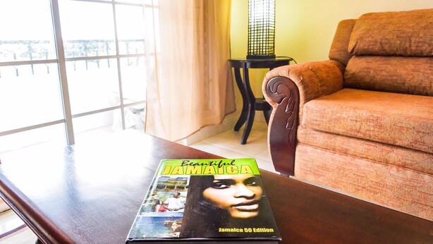 A living room with a sofa, a table with a book titled "Beautiful Jamaica 50 Edition," a lamp, and a window with curtains. - Kingston Guest Apt at Plantation Springs (Kingston)