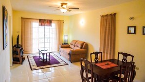 A living room with a sofa, a dining table with chairs, a ceiling fan, and a window with curtains. - Kingston Guest Apt at Plantation Springs (Kingston)