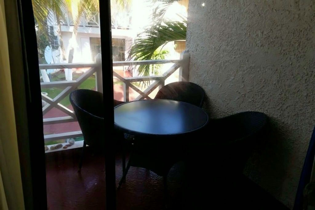 A small, enclosed patio area with a round table and two chairs, a white railing, and a view of a building and greenery outside.
