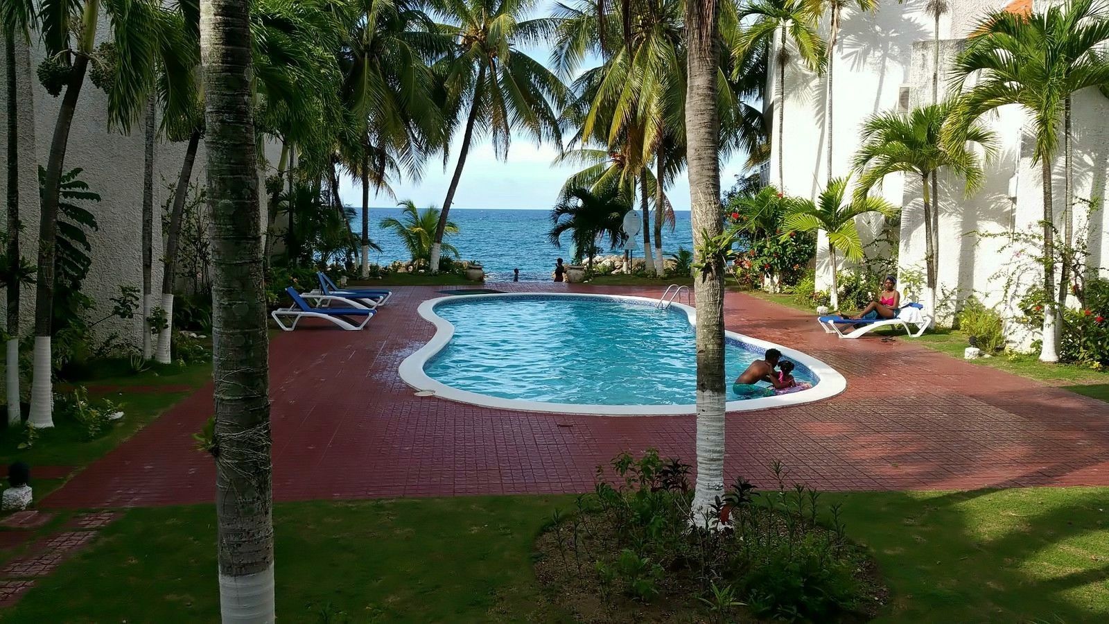 A pool area with palm trees, a red brick path, and a view of the ocean.