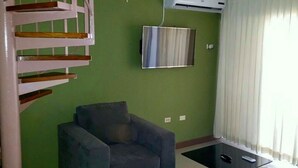 Een woonkamer met een grijze fauteuil, een flatscreen televisie aan de muur en een trap met houten treden.
