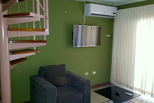 Una sala de estar con un sillón gris, un televisor de pantalla plana montado en la pared y una escalera con escalones de madera.