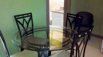 A round glass table with four chairs, a green wall, and a glimpse of a kitchen in the background.