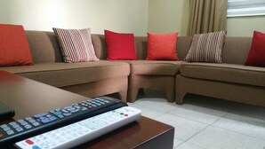 A living room with a sectional sofa, two remote controls on a coffee table, and a window with curtains. - Oaklands Guest Oasis (Kingston)