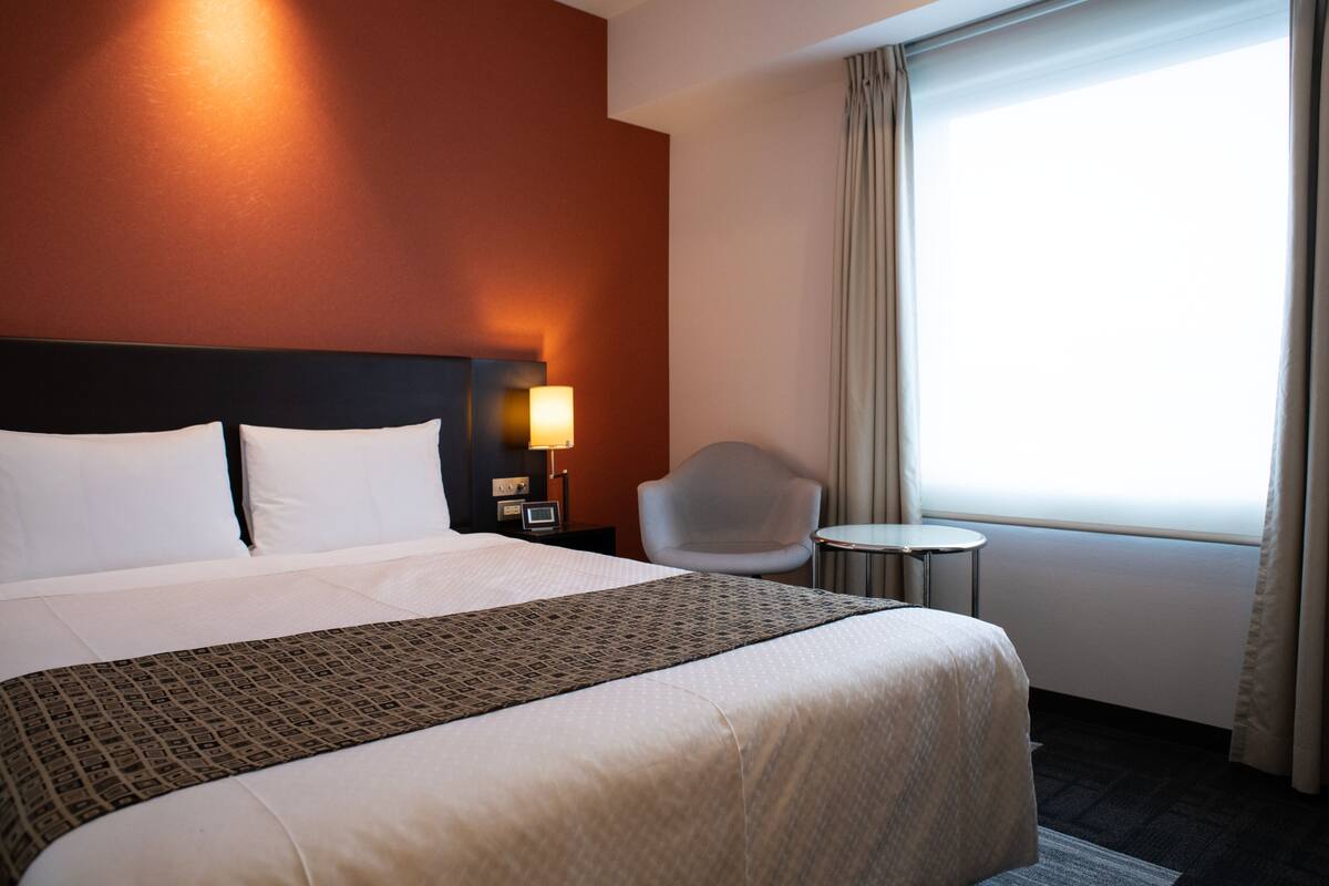 A hotel room with a large bed, a bedside table with a lamp, a chair, and a small table.