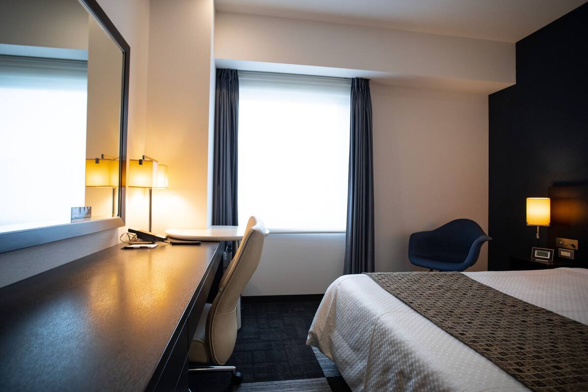 A hotel room with a bed, a desk with a chair, a lamp, a window with curtains, and a wall-mounted TV.