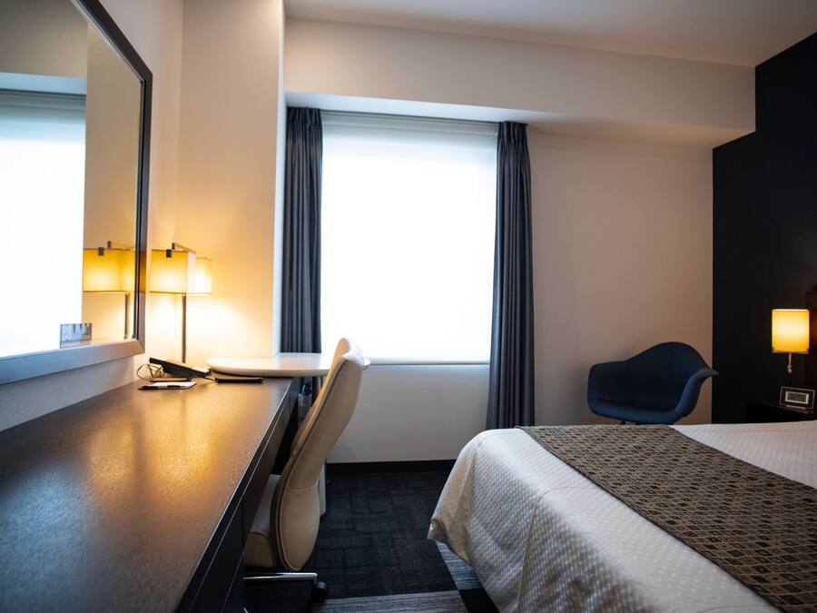 A hotel room with a bed, a desk with a chair, a lamp, a window with curtains, and a wall-mounted TV.