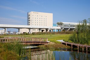 A modern hotel with a flat facade and multiple windows, a bridge over a pond, and a grassy area with a small water body. - Hotel Bestland (Tsukuba)