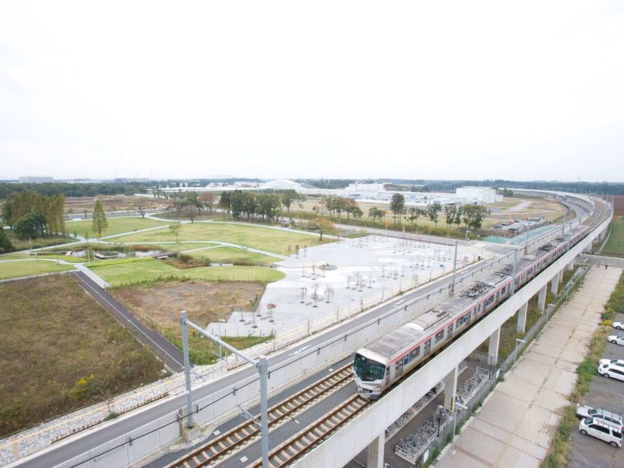A train station with a long elevated track, a parking area with cars, and a large building in the background.