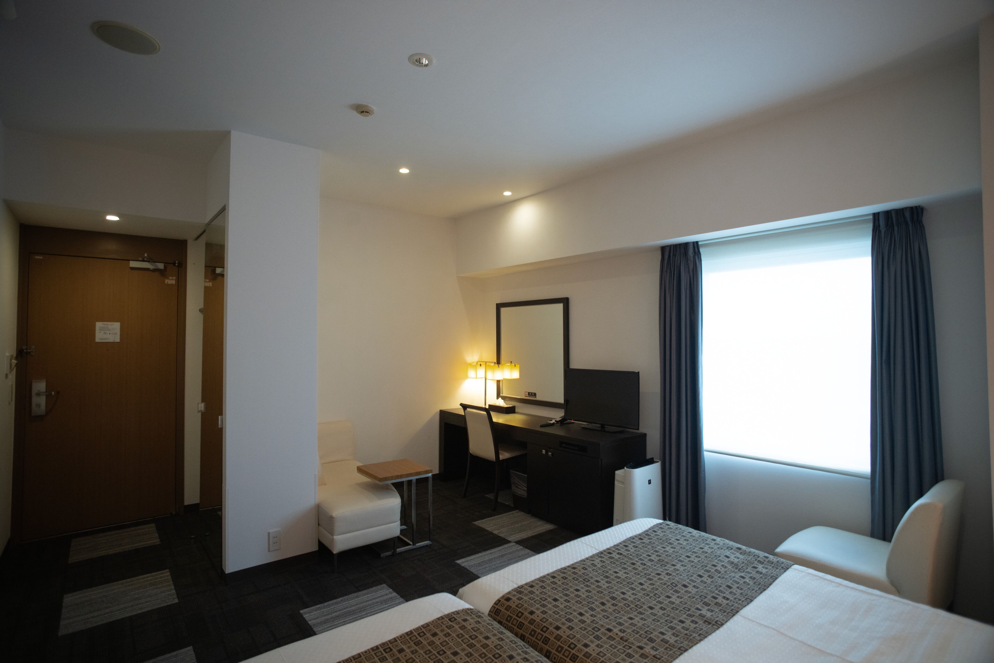 A hotel room with two beds, a desk with a chair, a TV, and a window with curtains.