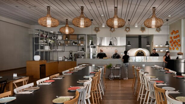 A restaurant with a long communal table, hanging wicker chandeliers, and a pizza oven in the background. - Jacob NEA  (Mateh Asher)