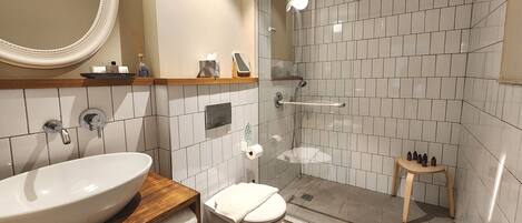 Un baño con un lavamanos grande y blanco, un inodoro, una ducha con puerta de vidrio y un estante de madera con artículos de tocador.