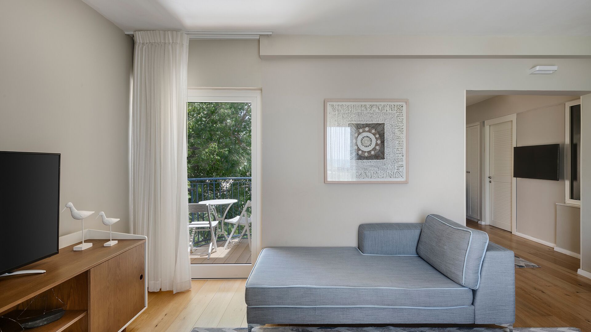 Un soggiorno moderno con TV a schermo piatto, un divano grigio, un'opera d'arte incorniciata e un balcone con vista su piante.