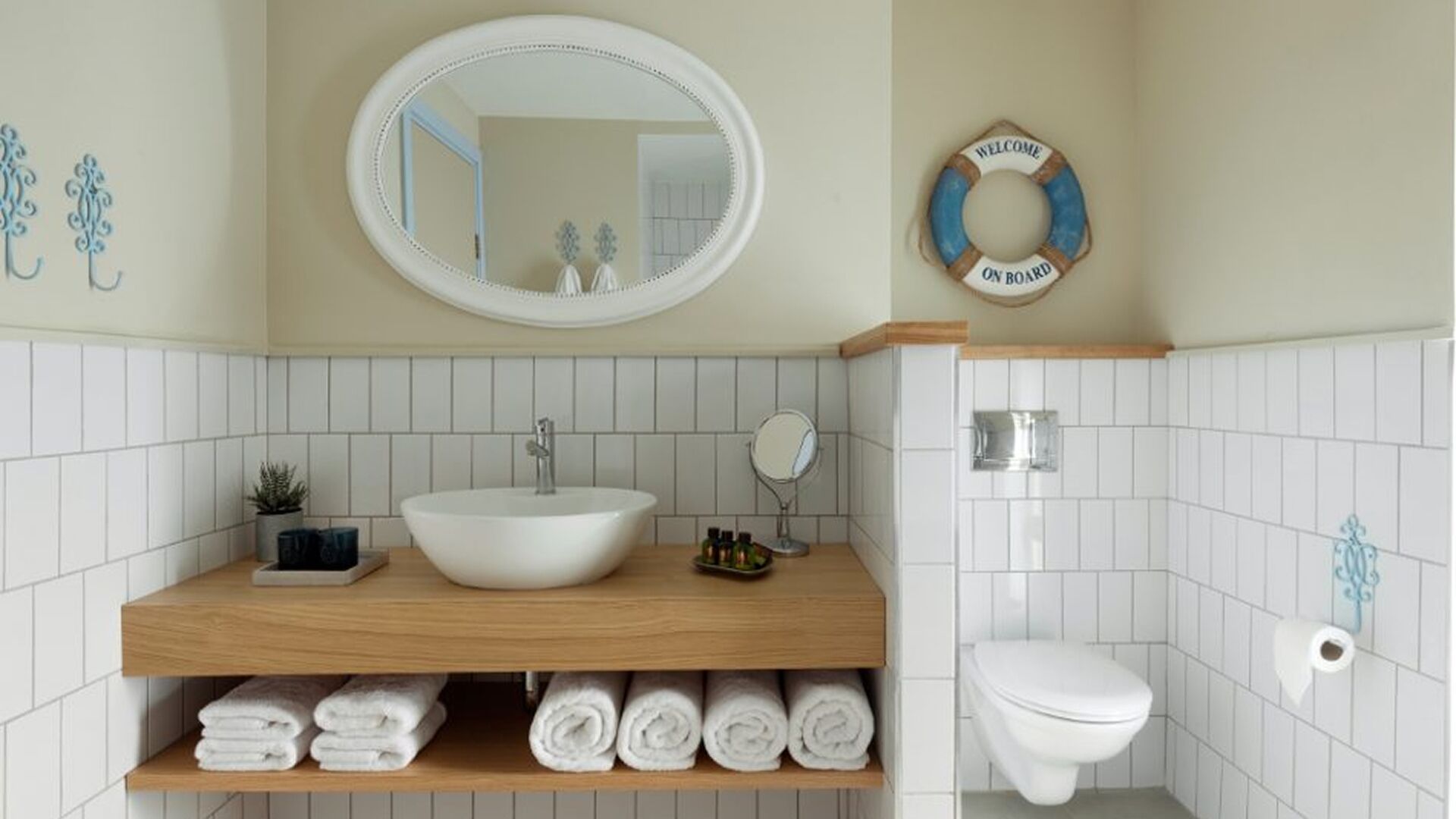 A bathroom with a white sink, mirror, and toilet, wooden shelves with towels, and a welcome sign.