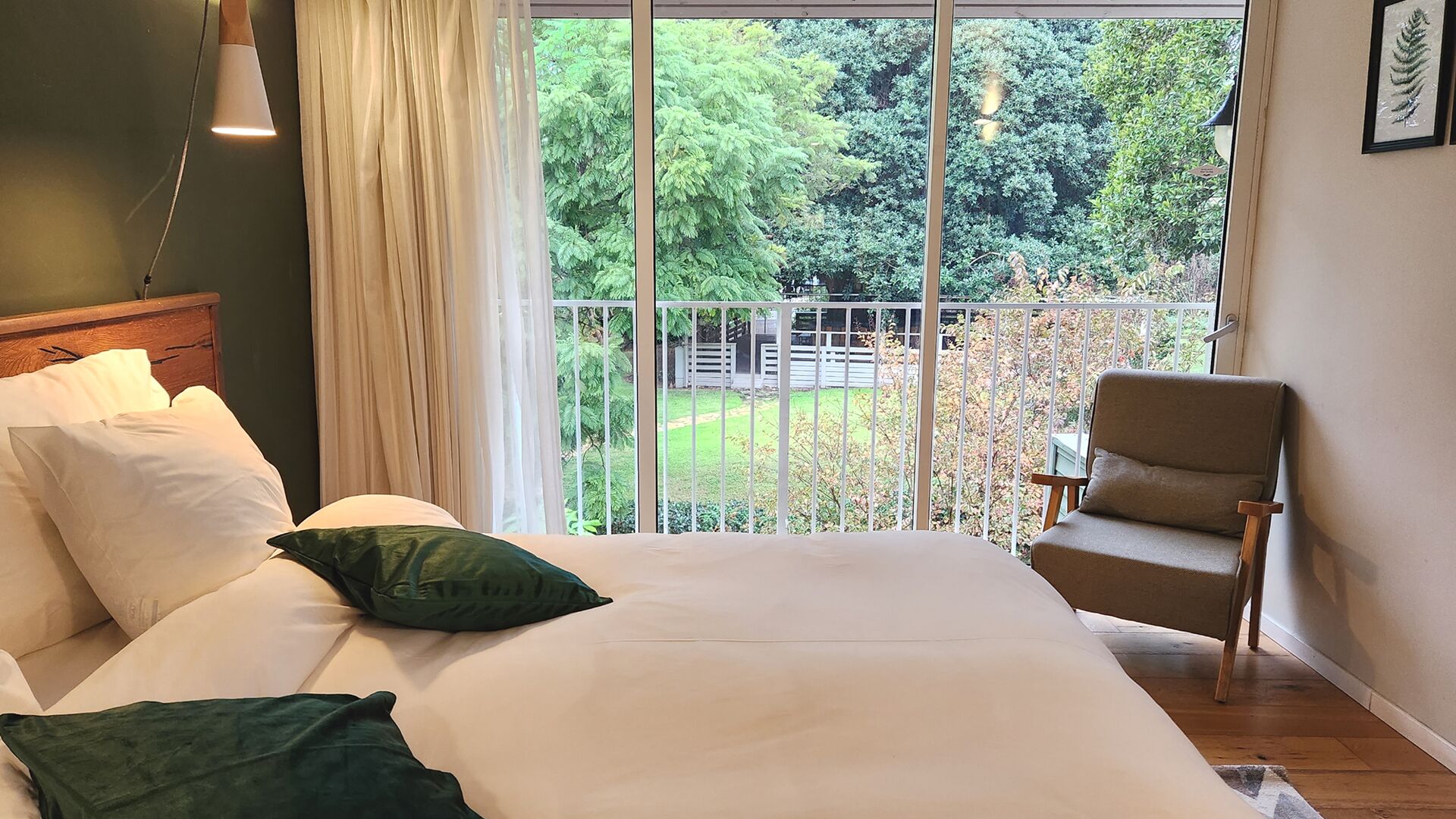 Un dormitorio con cama, almohadas, una silla, una ventana con vista a árboles y una barandilla de balcón.