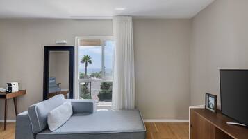 Habitación de hotel moderna con sofá gris, televisor, escritorio con cafetera y vista al mar a través de un ventanal amplio.