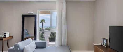 A modern hotel room with a grey sofa, a TV, a desk with a coffee maker, and a view of the sea through a large window.