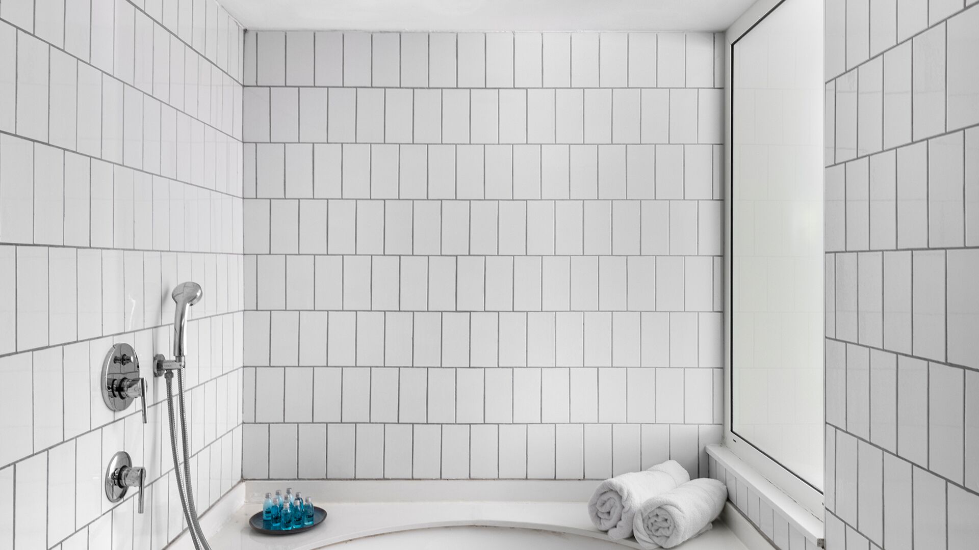 A modern bathroom with white subway tiles, a shower with a handheld showerhead, and two rolled towels on the floor.