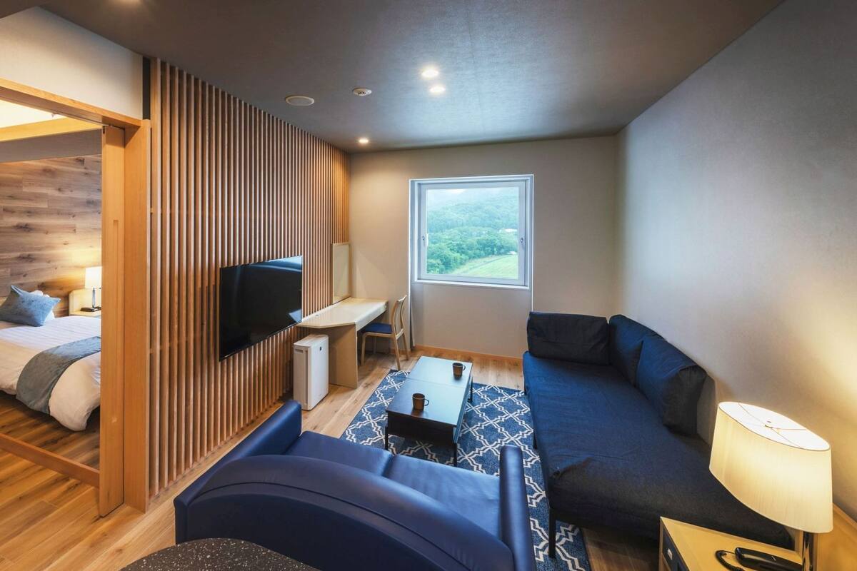 A modern hotel room with a bed, a TV, a sofa, a desk, and a window with a view.