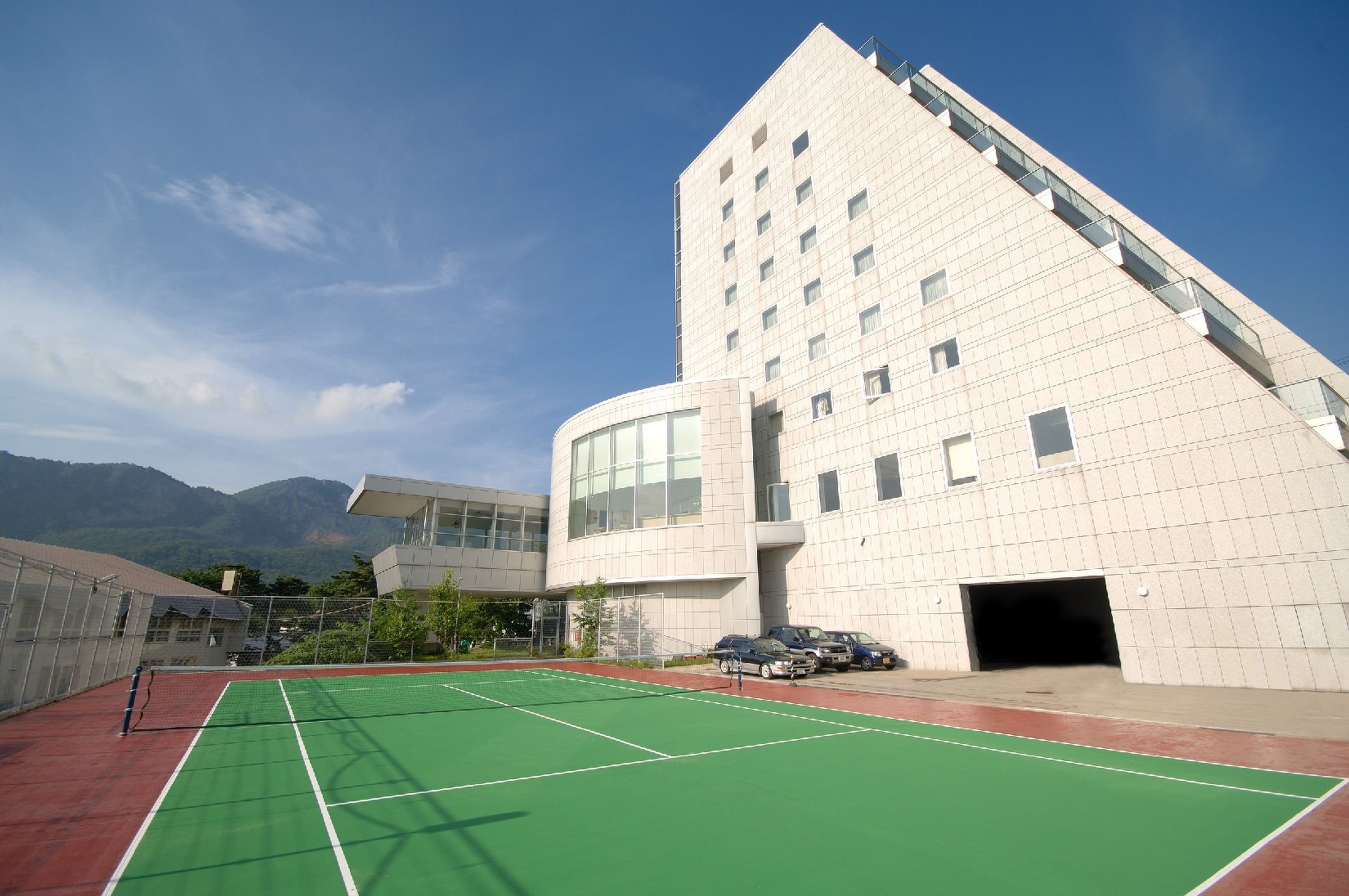 A modern building with a unique angular design, a tennis court in front, and mountains in the background.