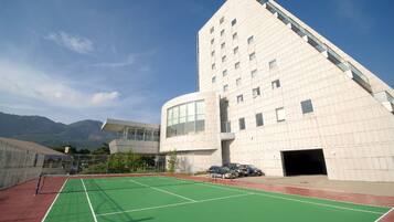 A modern building with a unique angular design, a tennis court in front, and mountains in the background.