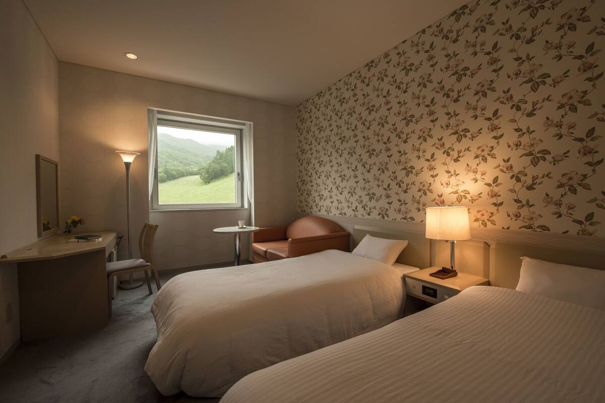A hotel room with two beds, a desk, a chair, a small table, and a window with a view of greenery.