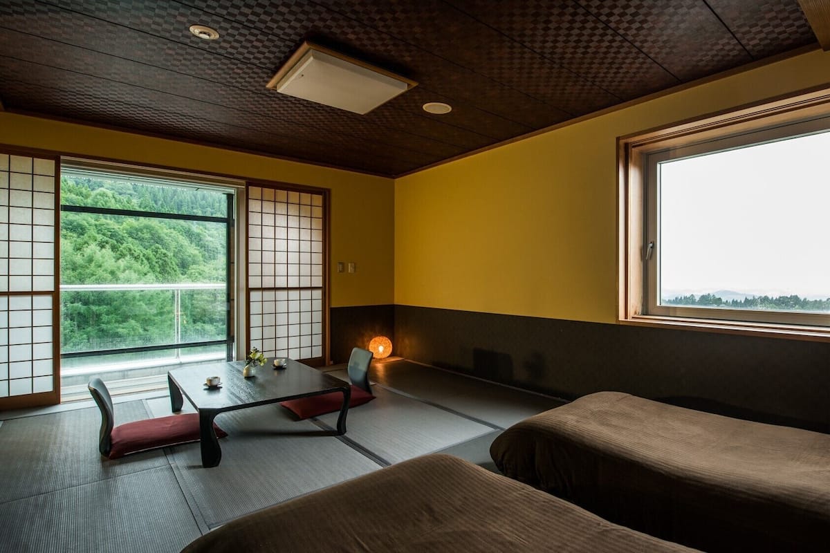 A room with tatami flooring, a low table with a cushion, a sofa, and a large window with a view of greenery.