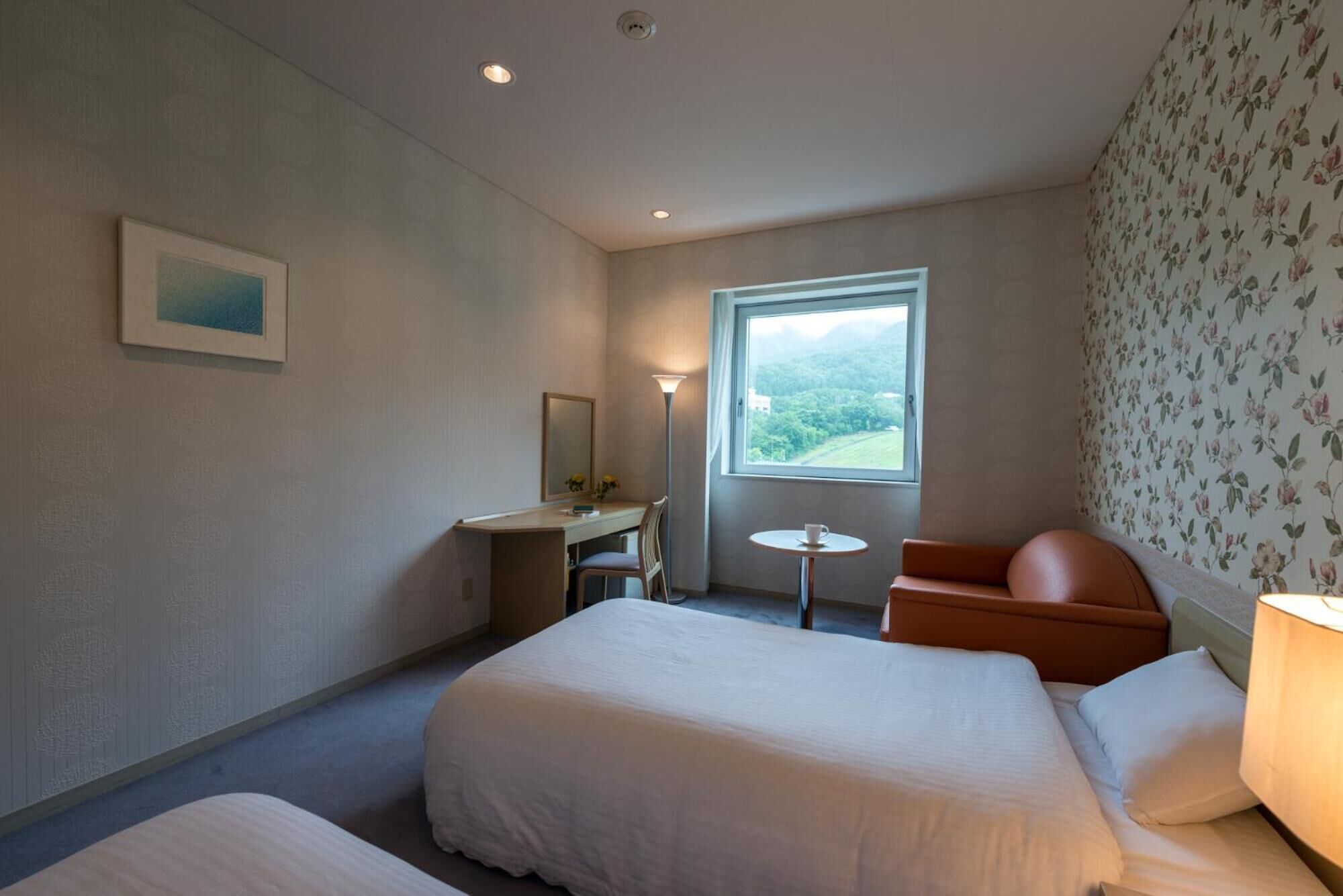 Habitación de hotel con dos camas, un escritorio, una silla, una mesita y una ventana con vistas a zonas verdes.