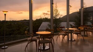 Une terrasse avec des tables et des chaises rondes, de grandes fenêtres et une vue sur un coucher de soleil se déclinant sur un paysage montagneux.
