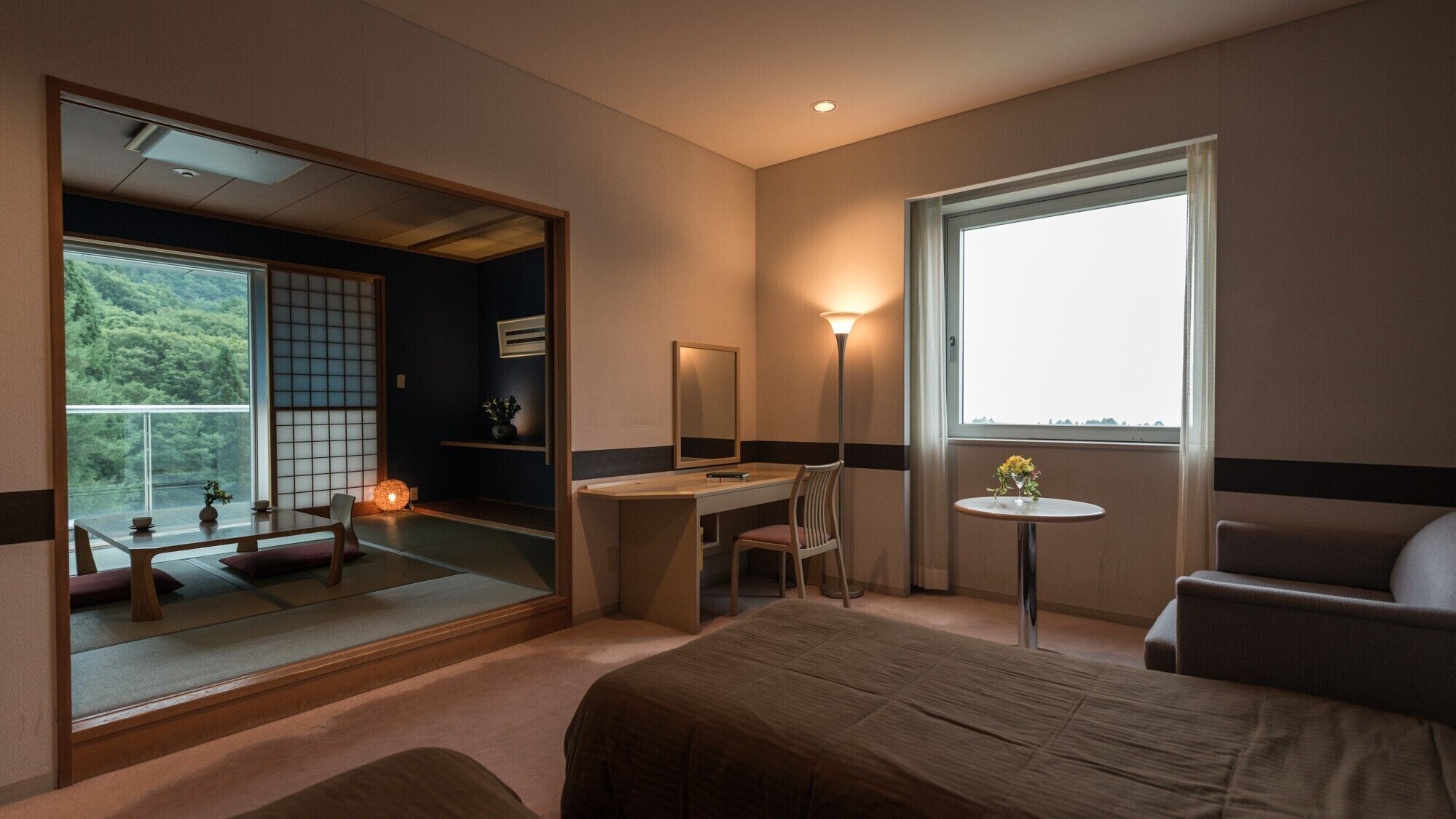 A hotel room with a bed, desk, chair, bedside table, and a large window with a view.