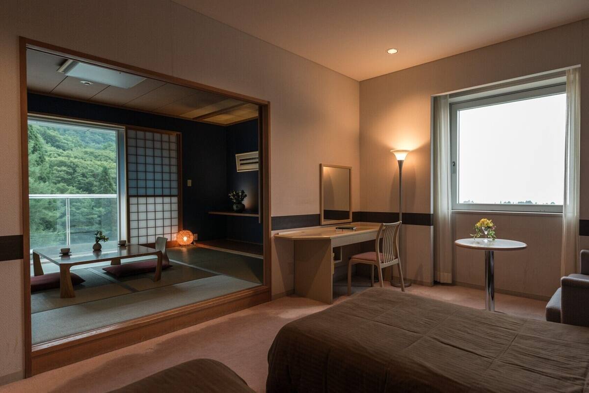 A hotel room with a bed, desk, chair, bedside table, and a large window with a view.