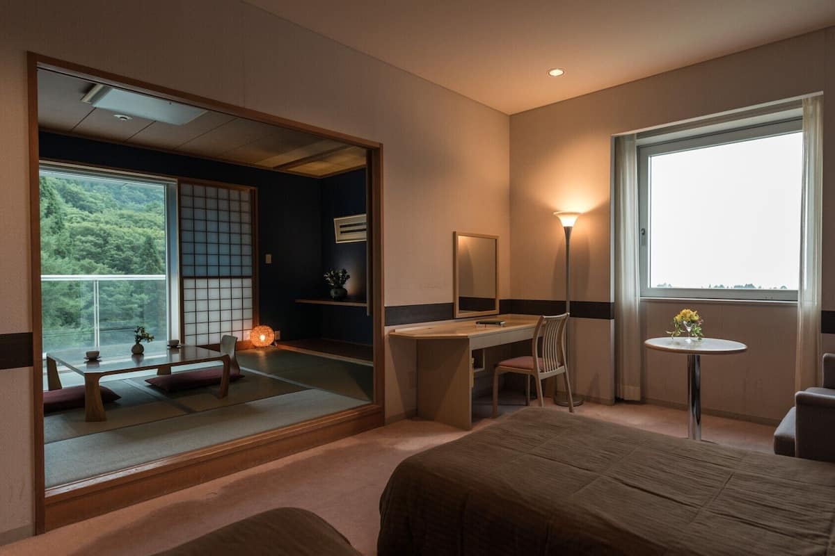 A hotel room with a bed, desk, chair, bedside table, and a large window with a view.