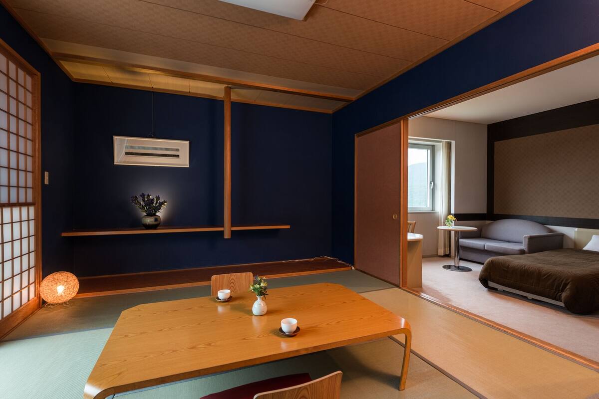 A traditional Japanese-style room with tatami flooring, a wooden table with a vase and a cup, and a sliding door leading to a living area with a sofa and a TV.