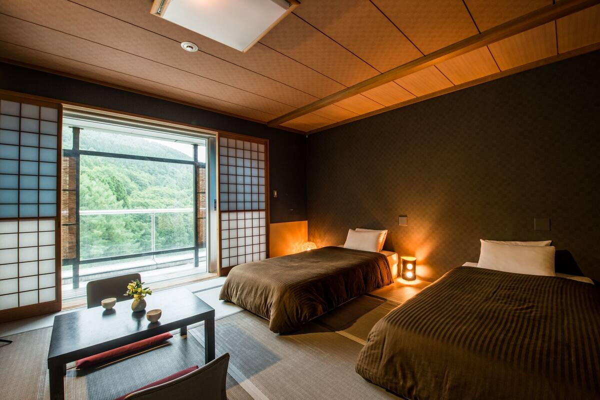A room with two beds, a table, and a view of greenery through large windows.