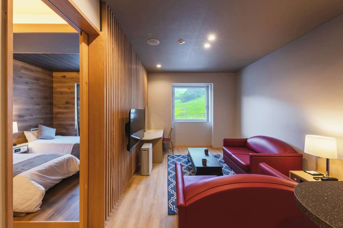 A compact room with a bed, a red sofa, a TV, a small table, and a window with a view of greenery.