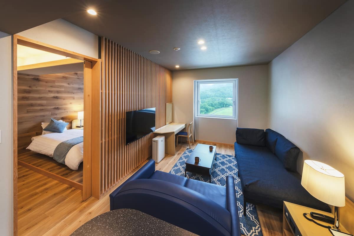 A compact hotel room with a bed, a sofa, a small table, a TV, and a window with a view of the outdoors.