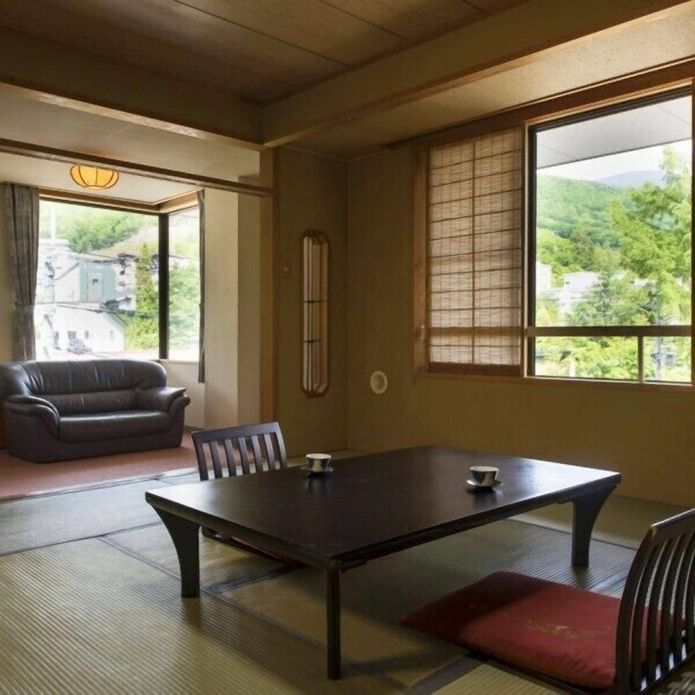 Um quarto em estilo japonês tradicional, com tatames, uma mesa baixa, um sofá e uma janela grande com vista para vegetação.