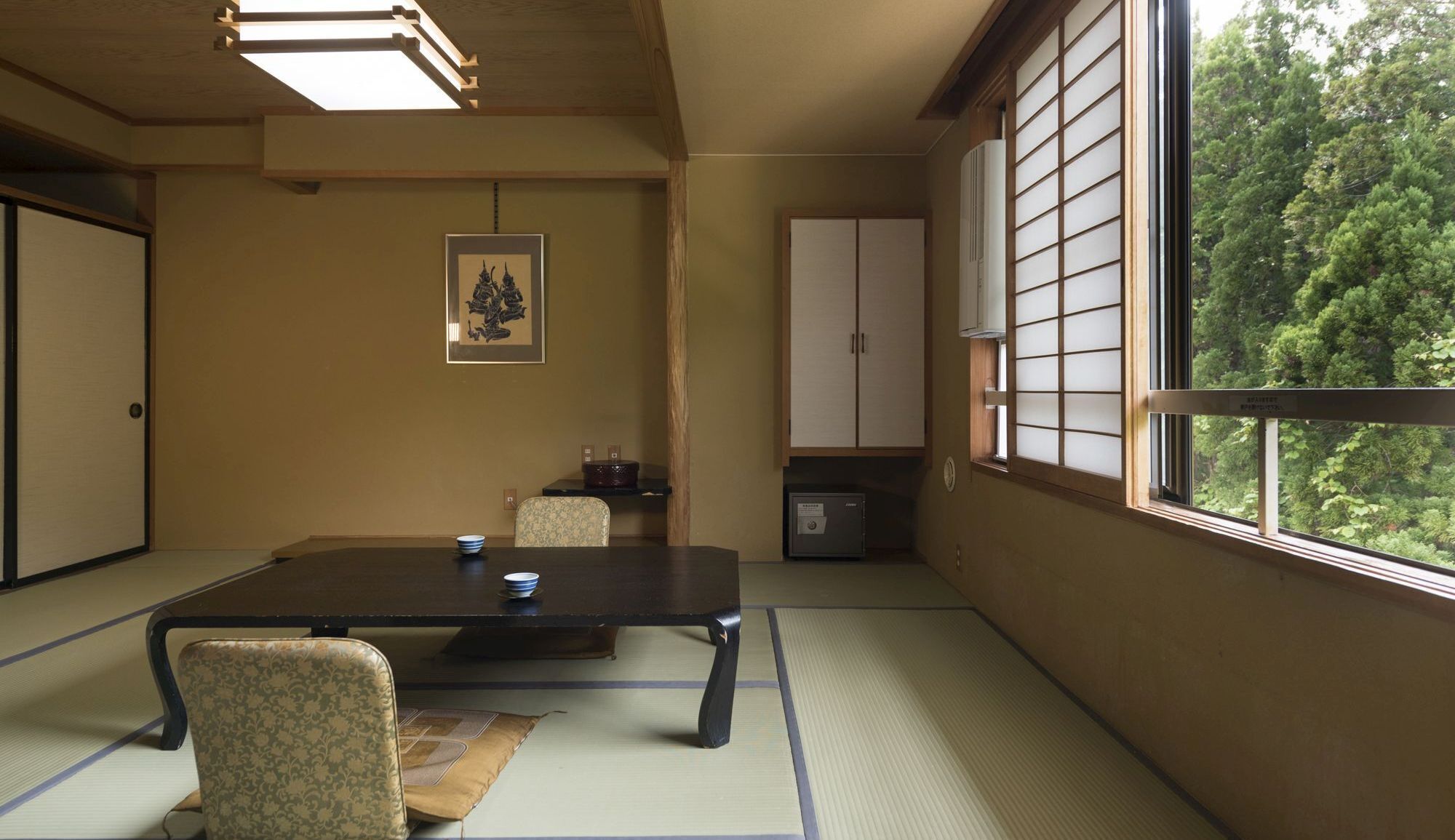 Una habitación con tatami, una mesa baja, sillas, un armario y un ventanal con vistas a árboles.