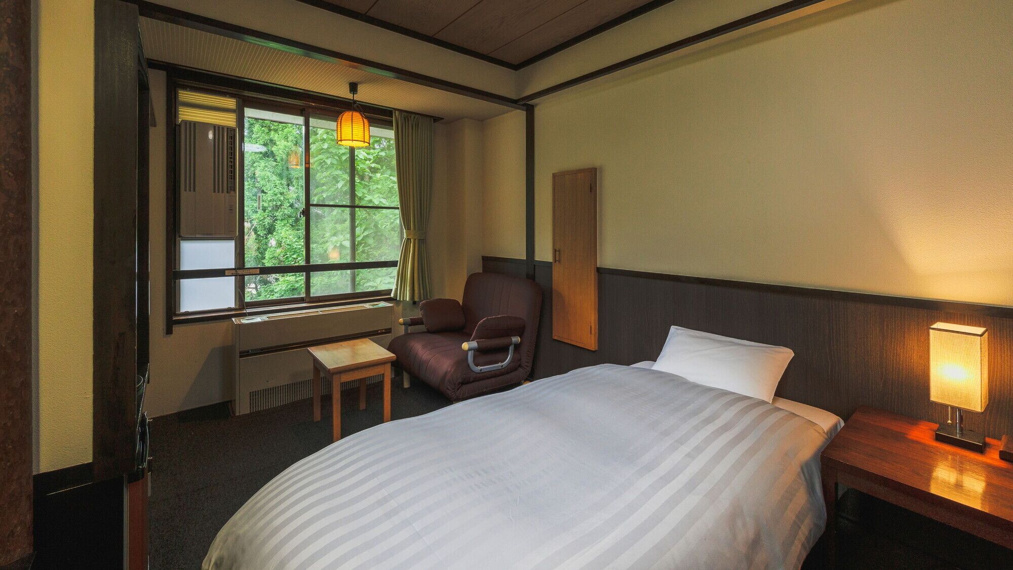 Habitación de hotel con cama, una silla, un escritorio y ventana con vistas a árboles.