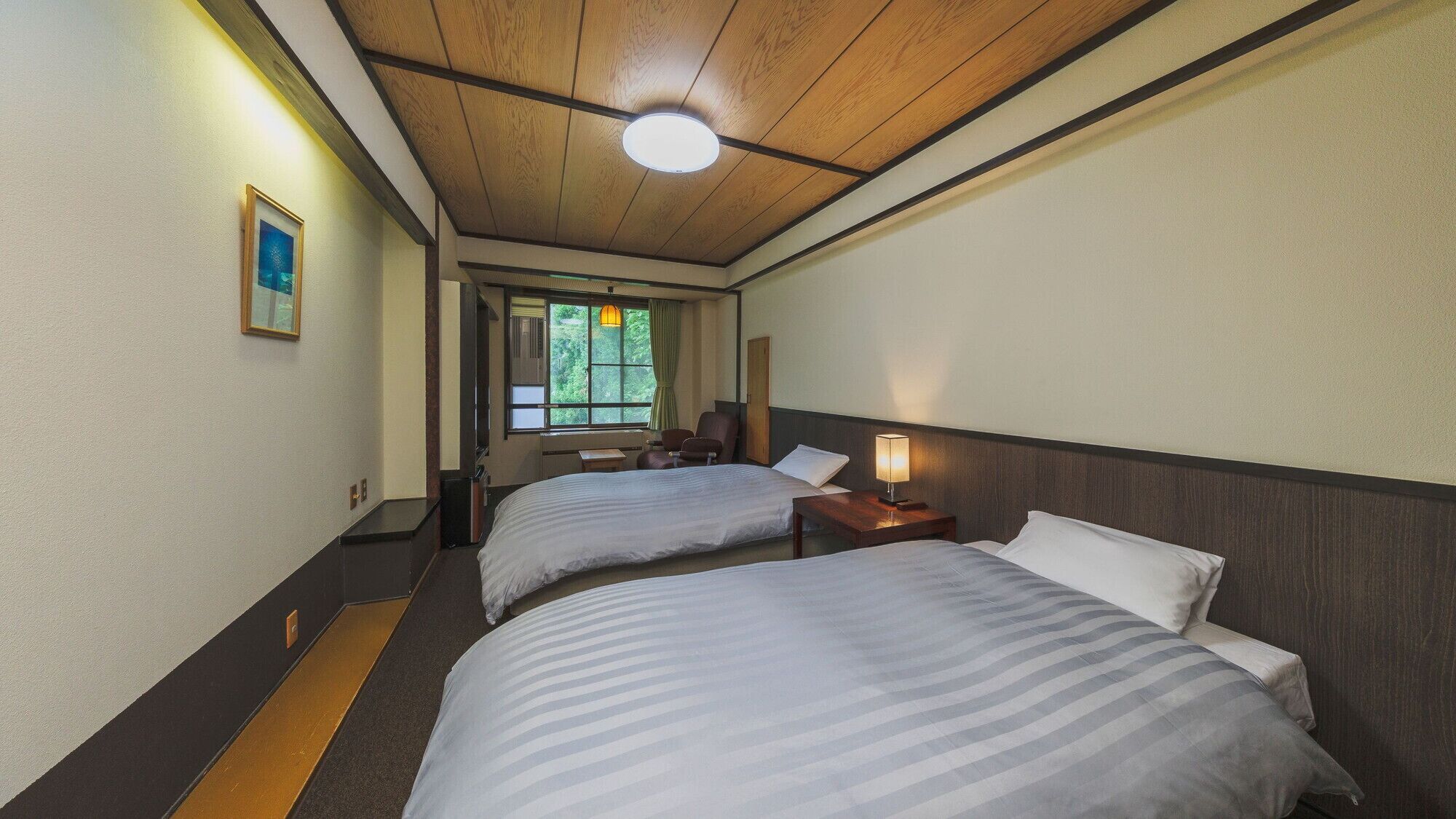 Quarto de hotel com duas camas, uma escrivaninha, uma cadeira, uma janela com cortinas e uma luminária no teto.
