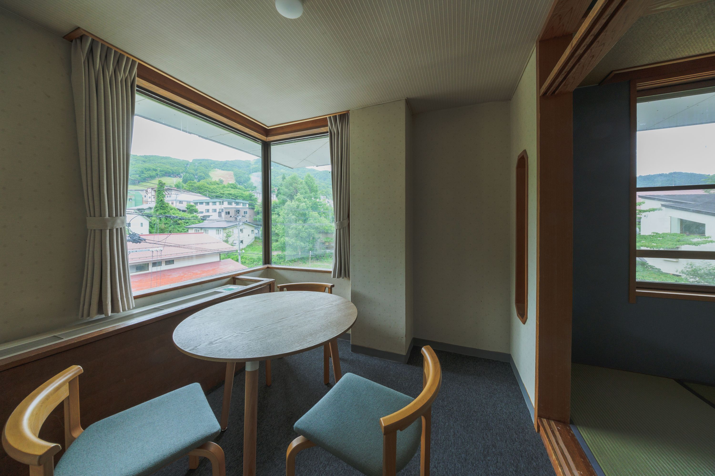 A room with a round table, two chairs, a large window with curtains, and a view of buildings and greenery.