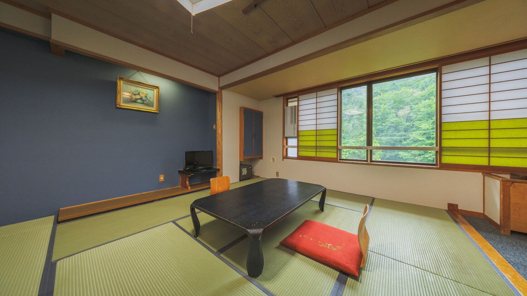 Um espaço com tatames, uma mesa de tatame, um assento vermelho, uma televisão e uma janela com biombos shoji.