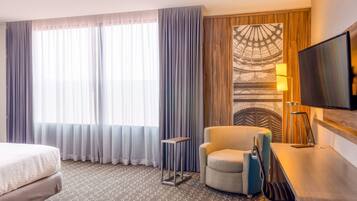 A hotel room with a bed, a chair, a desk, a TV, and a large window with curtains.