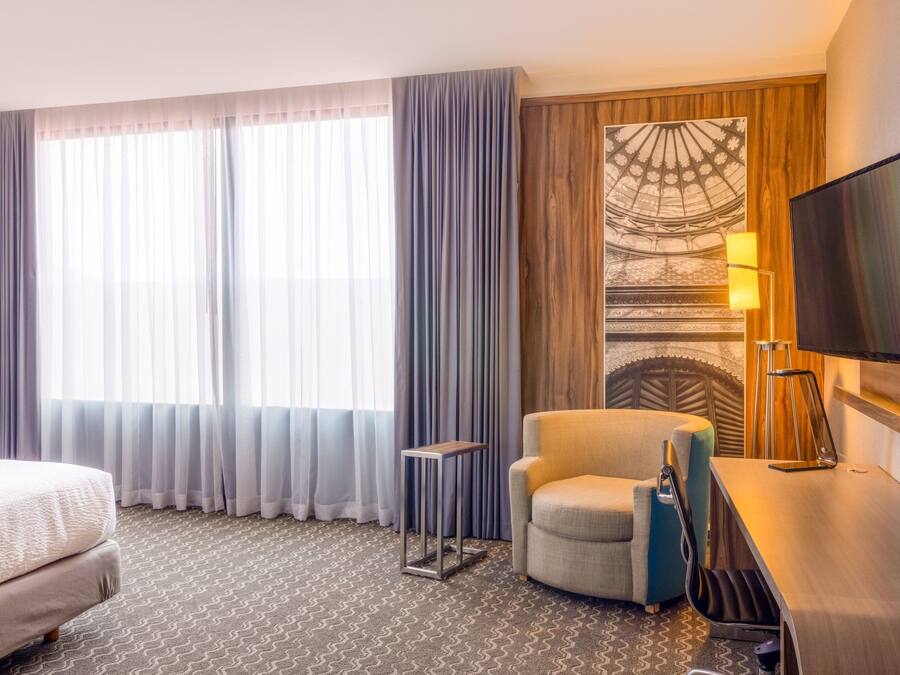 A hotel room with a bed, a chair, a desk, a TV, and a large window with curtains.